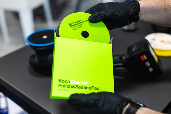 Person holding a Koch Chemie polish and sealing pad with gloves on, against a blurred background.
