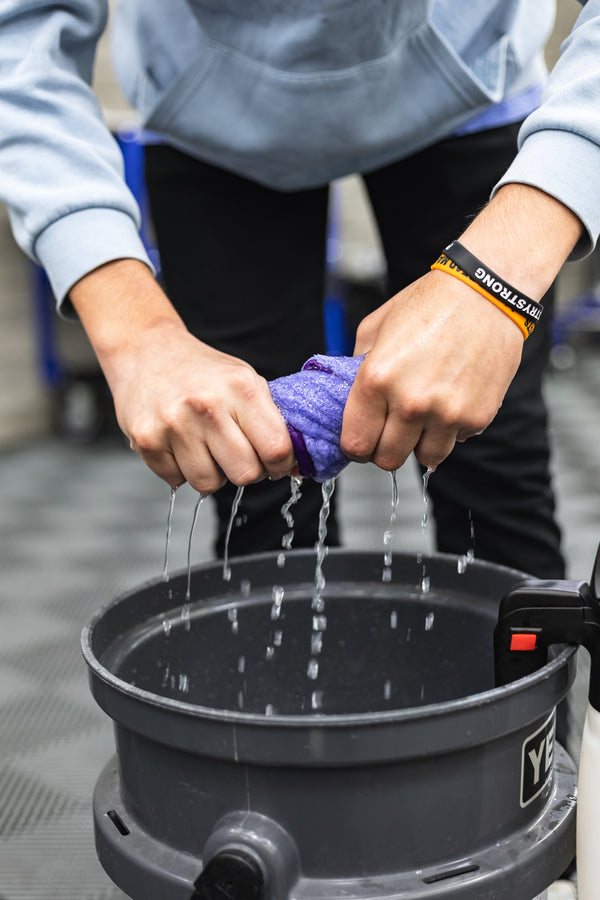 Person wringing out a purple minx royale towel into a black bucket with a YETI logo.