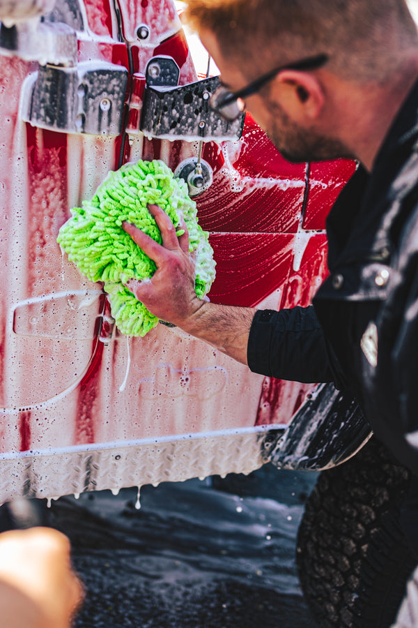 Person washing a red vehicle with a green microfiber wash mitt, focusing on the lower side panel, with the vehicle covered in soap suds.