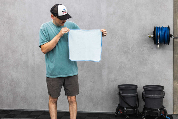 Person holding a blue ftw microfiber towel against a gray wall next to buckets and a hose.