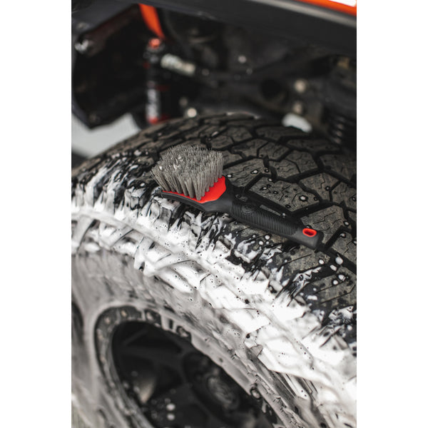 A red Detail Factory Tire Scrub Brush resting on a tire covered in foam.