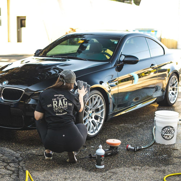 A person cleaning a black BMW coupe with a grey towel while wearing a rag company t-shirt.