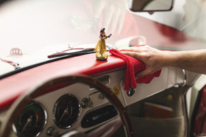 Hand cleaning the dashboard of a classic car with a red microfiber cloth, next to a small hula dancer figurine.