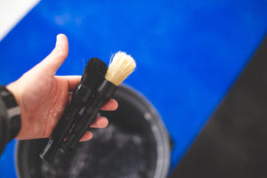 A person holding two Detail Factory black Boar Hair Brushes in front of a blue background.