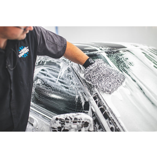 A closeup of someone using The Rag Company Ultra Wash Mitt on the front mirror of a black car covered in foam.
