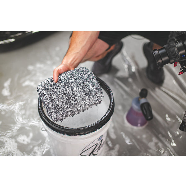 A closeup of someone holding The Rag Company Ultra Wash Mitt over a white bucket with a black rim.
